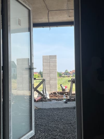 The image captures a view through an open doorway onto a construction site in Strumivka, Ukraine. In the foreground, a partially open white-framed window with clear glass reflects the sky and the surrounding area. The construction site itself features stacks of gray concrete blocks, one of which is a tall, narrow column. Adjacent to this is a wooden structure with diagonal supports. Several pieces of construction material, including pipes and a red-orange pipe, are visible. A bucket containing some red items and a gray pipe is also present. A plastic water bottle sits on a ledge near the bucket. In the background, beyond the immediate construction zone, several houses with pitched roofs and trees are visible under a clear blue sky. The ground in the foreground and mid-ground is covered with gravel. The overall scene depicts an active or recently active construction area.