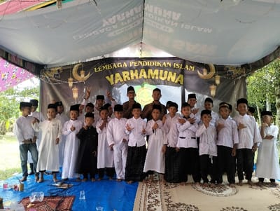 Under a temporary white canvas canopy, twenty-four males, predominantly young boys, are arranged in rows for a group photograph in Jambo Dalam, Indonesia. Most individuals wear white traditional Islamic attire, including long-sleeved shirts or tunics and dark trousers or sarongs, complemented by black kopyah (caps), some with gold trim. Two taller individuals in the back, possibly instructors, wear brown patterned shirts and kopyah.
The subjects stand on a covered ground; the left side is a blue tarp with scattered plastic bottles, cups, and electronic cables, while the right side features a patterned, light-colored rug. In the background, a large black banner displays "LEMBAGA PENDIDIKAN ISLAM YARHAMUNA" in yellow and white text, flanked by crescent moon and lantern motifs. Several boys are actively posing: one in the back makes a peace sign, another points a finger upwards, one in the front gives a thumbs-up, and others place hands over their chests or cross their arms. Beyond the canopy, green trees and foliage are visible under clear daylight, indicating an outdoor setting.