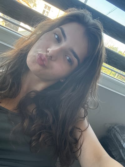 A young woman with long, dark, wavy hair is taking a selfie. She is wearing a dark green top. Her face is angled towards the camera, and she is puckering her lips. Her eyes are open, and she is looking directly at the camera. Her eyebrows are dark and well-defined. She has a small diamond earring in her visible ear.

The background suggests an indoor setting with blinds partially covering a window. Through the blinds, one can see sunlight and hints of trees and buildings, suggesting an urban or suburban environment. The lighting indicates it is daytime, with natural light coming from the window. No specific landmarks in Dibich, Bulgaria, can be identified from the image.