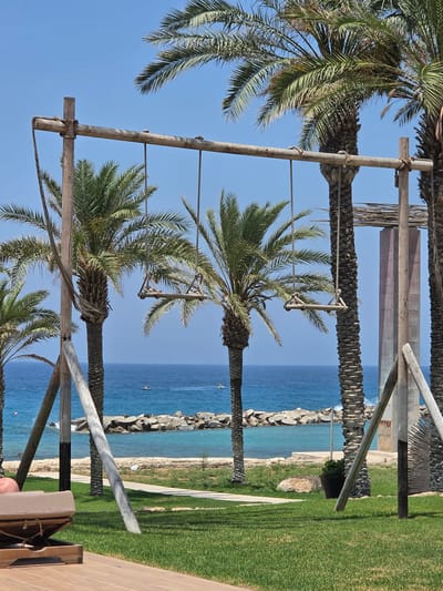 In Hlorakas Community, Cyprus, a tropical scene unfolds with a focus on a handmade wooden swing set, prominently featured in the foreground. The swing set, constructed from rough-hewn timber, supports two individual swings. Each swing consists of a wooden plank suspended by ropes. Surrounding the swing set are several tall palm trees, their fronds casting dappled shadows on the manicured green lawn.
Beyond the lawn, the cerulean expanse of the Mediterranean Sea stretches towards the horizon, dotted with two small white boats. A breakwater made of large grey rocks separates the sea from a tranquil turquoise bay. The shore is lined with sand and sparse vegetation. In the foreground, a portion of a wooden deck and a reclined lounge chair are visible, suggesting a resort or recreational area. The sky is a clear, bright blue, indicative of a sunny day. The overall ambiance is one of serene coastal relaxation.