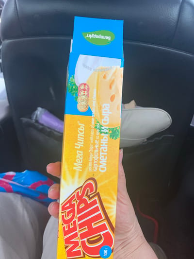 A person's hand is holding a bright yellow and blue box of "Mega Chips" potato chips with a sour cream and cheese flavor. The chips are packaged in a rectangular cardboard box. The box features an image of cheese and text in Russian, including "Мега Чипсы" (Mega Chips) and "сметана и сыр" (sour cream and cheese). The background appears to be the interior of a vehicle, possibly a car, with dark upholstery visible. The setting is indoors and the lighting suggests it is daytime. The location within Курапольскі сельскі Савет, Belarus is not discernible from the image.