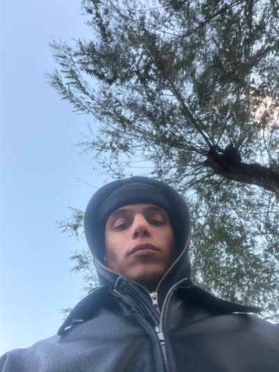 A low-angle outdoor image depicts a young individual in Migdal HaEmeq, Israel. The subject, positioned in the lower half of the frame, faces the camera directly with a neutral expression. They wear a black beanie, a dark hooded garment with the hood up, and a black leather-like jacket featuring prominent silver zippers. The upper portion of the scene consists of a pale blue sky on the left, and a tree with dense green foliage and visible branches on the right, extending across the upper background. No other subjects or objects are discernible.