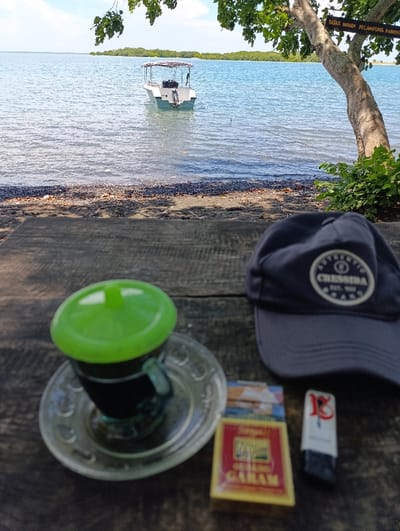The image shows a wooden table set up on a beach, overlooking a body of water in Kecamatan Banyuputih, Indonesia. There is a small boat in the distance. A cup of coffee with a green lid sits on a glass saucer. A pack of cigarettes and a lighter are on the table next to the cup. A black baseball cap with a logo reading "CRESNIDA" is lying on the table to the right. The camera is positioned on the table and angled towards the water. The scene is calm and peaceful. The tree in the background creates shade on the table and on the sand. The beach is mostly black sand with a few patches of white sand. The water is calm and clear. The boat is a white, flat-bottomed vessel with an outboard motor. The sun is shining, and the sky is blue. There are no people visible in the image.