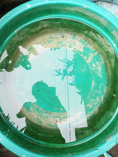 A circular teal-green plastic tub, exhibiting internal wear and yellowish-green staining, is partially filled with clear water. The water's surface acts as a mirror, reflecting the overhead environment and a human figure. A distinct silhouette of an individual, presumed to be the photographer, is prominently reflected in the lower-central portion of the water, holding a device. Reflections of green foliage are discernible along the left perimeter of the water, while vertical and diagonal lines, indicative of overhead utility wires or structural elements, are reflected in the upper-right quadrant. The scene is located in the city of Pandaria, India.