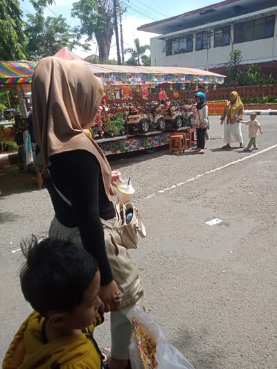 The scene depicts a sunny outdoor environment in Nganjuk, Indonesia, specifically a paved street adjacent to a vendor area. In the immediate foreground, a person wearing a light brown hijab, black long-sleeved shirt, and light-colored pants stands facing right, holding a clear plastic cup with a beverage and straw in the right hand. A light-colored shoulder bag hangs from their left side. A young child with dark hair, wearing a yellow hoodie, stands to the adult's left, looking right, holding a clear plastic bag containing colorful confectionery. The adult's left hand appears to be holding the child's arm or hand.
In the midground, a vibrant street vendor stall is visible, featuring a rainbow-striped canopy and displaying multiple child-sized ride-on vehicles (miniature cars and ATVs) along with various plush items. Two small brown stools are positioned near the stall. To the right of the stall, on the asphalt street, three individuals are present: one person wearing a blue helmet and light attire, standing near the stall; a woman in a yellow hijab and white dress, holding the hand of a small child; and the child, dressed in light-colored clothing, walking alongside the woman. A yellow sign with red lettering partially reading "DILARANG PARKIR PENGGALAN" (No Partial Parking) is visible behind them.
The background features a multi-story building with windows, exhibiting a reddish-brown lower facade and a lighter upper section. Numerous green trees and foliage are present, along with utility poles and overhead wires. The sky is clear blue with minimal cloud cover.