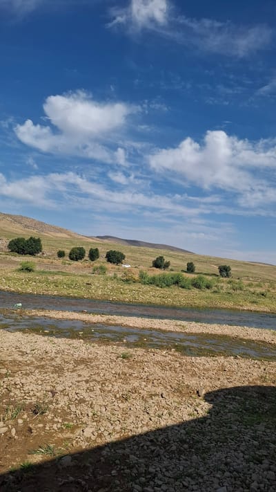 The image depicts a wide, shallow river flowing through a rocky, arid landscape. The riverbanks are covered in pebbles and sparse vegetation. In the middle ground, a gentle, grassy hillside rises, dotted with scattered trees and a white vehicle. The background shows rolling hills under a bright blue sky with scattered white clouds. The scene appears to be a natural, rural environment.