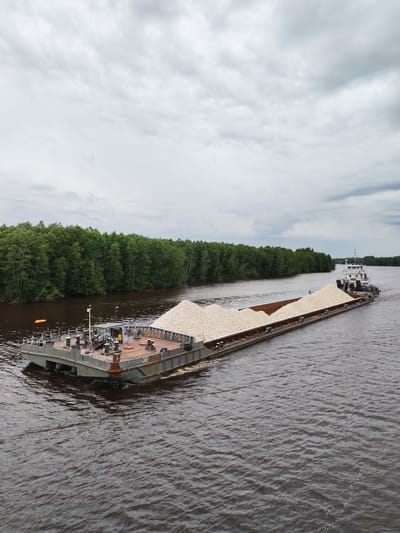 A barge laden with a large quantity of light-colored, granular material, possibly aggregates or minerals, is being towed on a wide, calm river. The barge, gray and utilitarian, has a visible deck with various equipment, including winches and bollards. A smaller tugboat is pushing the barge from the rear. The river is dark brown, suggesting it's carrying sediment.
On the left bank of the river, a dense, green forest lines the water's edge, with tall trees creating a thick canopy. The sky is overcast with heavy, gray clouds, indicating a possibly cool and damp day. The overall scene depicts industrial transportation in a natural setting, likely during daytime.
On the barge, visible text can be partially deciphered as "N 8157" and possibly some Cyrillic letters. The environment suggests a location in Russia, given the mention of Novokemskii. The mood is one of steady, industrial activity, juxtaposed with the serene natural landscape.