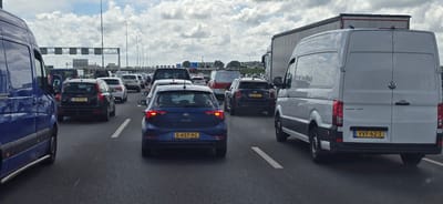 The image shows a traffic jam on a multi-lane highway, likely the A4 motorway, near Hoofddorp, Netherlands. Several vehicles are stationary or moving slowly. The sky is cloudy.

In the foreground, on the left, is the rear of a blue van. To the right of the van, a dark Volvo V40 station wagon with license plate "33-XZ-KL" is visible. Following it is a dark blue Volkswagen Polo with license plate "S-457-XG". Further ahead, there is a black Mini Cooper with license plate "RD-586-F" and several other cars and vans. A large white Volkswagen Crafter van with license plate "VVF-62-Z" is positioned in the right lane. A large articulated truck is also present in the background. Traffic lights and overhead signs for road directions are visible above the lanes. The road surface appears wet, suggesting recent rain. There are no people clearly visible in the image. The scene depicts a typical rush hour traffic congestion.