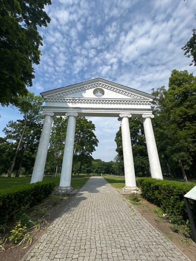 The image shows a low-angle, full shot of a grand white stone archway in a park setting. The archway features a triangular pediment with decorative molding and a circular emblem at its apex. Four large, white cylindrical columns, two on each side, support the arch. The columns appear to be leaning slightly inwards. A paved pathway, made of grey cobblestones, leads directly towards the archway. To the left and right of the pathway are well-maintained green bushes. Beyond the archway, the park extends with more greenery and tall trees under a bright blue sky with scattered white clouds. The scene suggests a sunny day in a public park. This landmark is known as the Jurbarkas Great Arch, a historical monument located in the city of Jurbarkas, Lithuania.