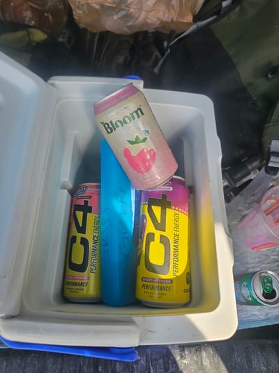 The image shows the interior of a cooler. Inside, there are cans of drinks stacked on top of each other. There is a pink "Bloom" brand can of strawberry cream drink, as well as two cans of "C4 Performance Energy" drink in yellow and red watermelon flavor. There is a blue ice pack lying in between the drinks with the name "Griff" written on it. In the top-left corner of the image, there is a green bag, and in the top-right corner, there is an open can of espresso. This image was taken in Big Pine Key, United States.