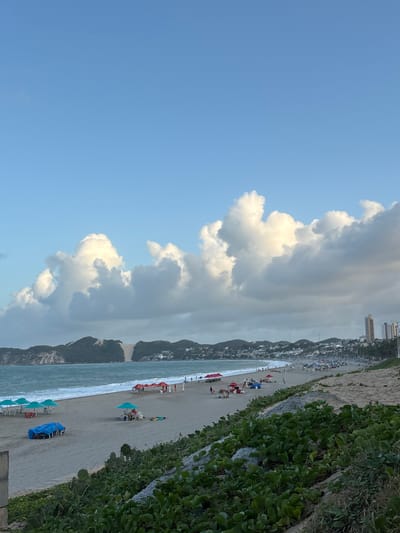 The image captures a broad view of Ponta Negra beach in Natal, Brazil. The sky is a light blue, partially covered by large, white cumulus clouds. The Atlantic Ocean's blue-green water, featuring white-crested waves, meets a wide, light-colored sandy beach. Along the beach, numerous individuals are visible, many congregating under various colored beach umbrellas, predominantly red, light blue, and dark blue. Some people are near the water's edge, while others are seated under umbrellas with chairs.
In the mid-ground, on the left side, the prominent Morro do Careca, a large sand dune, rises from a forested hillside. The coastline extends to the right, revealing further developed areas with additional green hills and several multi-story buildings in the distant background. The immediate foreground consists of a sandy slope covered densely with dark green, low-lying coastal vegetation. A light-colored structural element, possibly concrete, is partially visible on the far-left foreground edge.