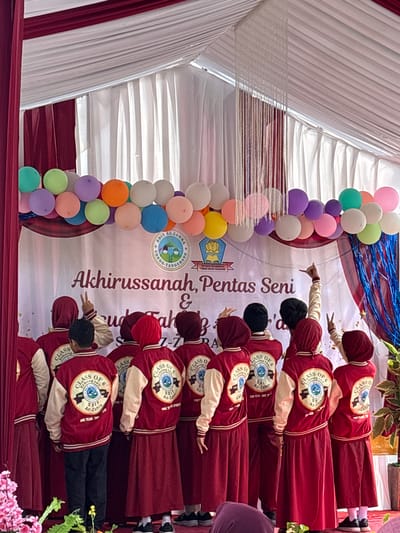 In Sukabakti, Indonesia, a group of students, primarily wearing maroon hijab and matching skirts, stand facing away from the camera. They are adorned with red varsity jackets featuring cream-colored sleeves and embroidered circular logos on the back, which read "CLASS OF 6" and "SDIT AZ-ZAHRA". Some students are raising their hands in the air, with one making a peace sign and another forming a lasso gesture. A banner behind them reads "Akhirussanah, Pentas Seni & Yudisium Fakh... 2027-2023", indicating a graduation and arts performance event. The backdrop is a draped white and maroon fabric, and a cluster of colorful balloons, including green, yellow, purple, orange, and pink, floats above. Decorative white string fringe hangs from the ceiling structure. The setting appears to be an indoor or covered outdoor stage.