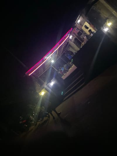The image captures a dimly lit street scene at night. Buildings are faintly visible in the background, their details obscured by darkness. A row of red and white lights stretches diagonally across the frame, possibly illuminating a structure or feature. Plants and parked motorbikes are clustered in the foreground to the right. Overall, the scene is dark and somewhat abstract, with the bright lights providing the primary visual interest.