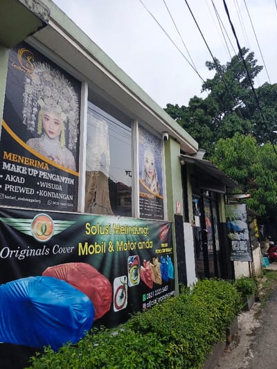 The image shows the exterior of a building with large window advertisements. "Lestari S" is advertised for makeup services, specifically for brides, graduations, pre-wedding shoots, and receptions. A second banner promotes "Originals Cover" car and motorcycle covers as a solution for protection. The banner displays images of covered cars and motorcycles in various colors, along with a phone number and social media handle. The building is light green, surrounded by green bushes, and has a view of trees and power lines in the background.