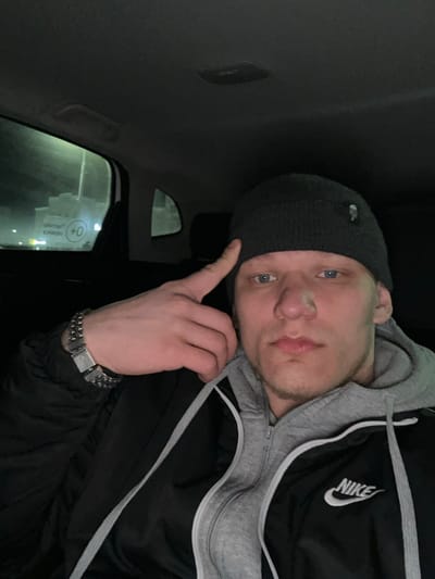 A young adult male is photographed in a vehicle interior at night in Dobryanka, Russia. The subject has light skin, blue eyes, light stubble on his chin, and a small white substance on the bridge of his nose. He wears a dark grey knitted beanie with a small dark label, a grey zippered hooded sweatshirt, and a black Nike brand athletic jacket with a white logo on the left chest. A wide, metallic link bracelet is present on his left wrist. His left hand is raised, with the index finger touching the beanie near his left temple. The vehicle interior is dark, showing the roof lining and a grab handle above the left rear window. Through this window, a blurred exterior is visible with artificial lighting and a sign displaying Cyrillic text, including "образов." and the prominent "0+" age restriction symbol.