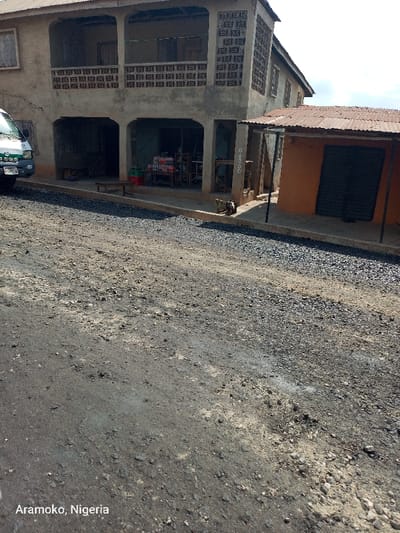 The image shows a street in Ilobu, Nigeria, at coordinates 7.836 latitude and 4.483 longitude. The street is paved with asphalt, which is cracked and uneven. There is a two-story building on the left side of the street and a single-story building on the right. The buildings appear to be shops, with a variety of goods visible through the windows. There is a white van parked on the left side of the street. There are no people in the image. The sky is clear. The image is taken from a street-level perspective.