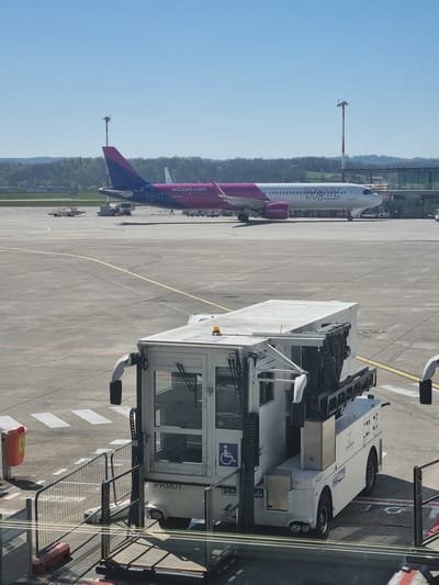 The image depicts an airport scene. A Wizz Air airplane is visible in the background, parked on the tarmac. A large white vehicle, possibly a passenger boarding bridge or service vehicle, is positioned in the foreground. The location appears to be outside an airport terminal. The city is Saint-Louis, France. There are no people visible in the image. The overall environment is sunny and bright.