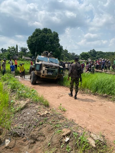 In Ayati, Nigeria, a rural or semi-rural landscape features a camouflaged armored personnel carrier (APC) or mine-resistant ambush-protected (MRAP) vehicle positioned on a muddy, unpaved dirt road under a partly cloudy sky. The vehicle, a 4x4 or 6x6, is primarily tan and green, with multiple windows and an open top hatch revealing at least one occupant. The front of the vehicle is submerged in a prominent mud puddle.
In the immediate foreground, a uniformed soldier, seen from behind, walks along the dirt road towards the vehicle and the crowd. The soldier wears dark camouflage fatigues and black boots, carrying a rifle.
Numerous civilians, predominantly men and boys, are gathered on both sides of the road, observing the scene. On the left, a smaller group of men and boys stands near the vehicle, some in the muddy area, with one boy in a yellow shirt holding his hands on his head. On the right, a larger crowd of civilians is assembled on an elevated patch of ground bordered by tall green vegetation.
The environment includes dense green foliage, tall grasses, and various trees, including a large deciduous tree in the background. Several traditional huts with thatched roofs are visible among the trees in the distant background. The dirt road itself is reddish-brown, with visible ruts and muddy sections.
