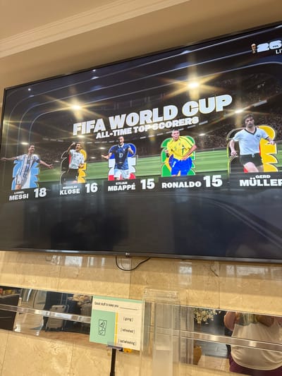 A large television screen displays "FIFA WORLD CUP ALL-TIME TOP SCORERS" with images of five soccer players. From left to right, the players are Lionel Messi (18 goals), Miroslav Klose (16 goals), Kylian Mbappé (15 goals), Ronaldo (15 goals), and Gerd Müller. The screen is mounted on a wall with a beige marble tiled background. Below the screen, a reflective surface runs horizontally, and further down, a stand with a sign that reads "Good stuff to keep you" with options "[going]", "[refreshed]", and "[cartwheeling]". A clear plastic dispenser is to the left of the stand. The overall scene appears to be in an indoor public space, possibly a lobby or waiting area, with reflections of luggage visible in the mirrored surface. The city of North Bergen, United States of America is not explicitly mentioned or visible in the image, but the presence of a FIFA World Cup display suggests a location with an interest in soccer.
