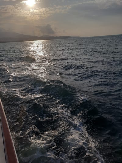 The image depicts a view from a boat, looking out at the Black Sea towards the distant coastline of Samsun, Turkey. The sun is partially obscured by clouds, casting a shimmering reflection on the water's surface. Gentle waves create a foamy wake trailing from the boat, visible on the right side of the frame, where the edge of the vessel also appears. The sky is overcast with scattered clouds, and the horizon is marked by a low, hilly coastline with faint outlines of mountains in the distance. The overall scene is one of serene, natural beauty at sea.