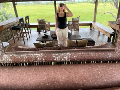 A person takes a selfie in a mirror in Khwai, Botswana. The person is a light-skinned female wearing a black tank top and beige pants. She holds a phone in front of her face. The reflection shows a covered patio area with wooden floors, chairs, and tables. Beyond the patio is a grassy field.