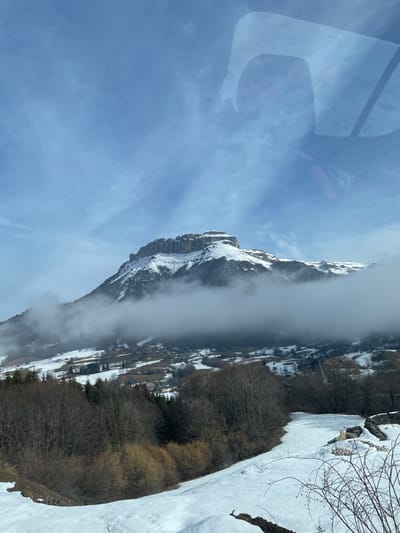 The image displays a winter mountain landscape in Les Déserts, France, viewed from an elevated perspective, possibly through a window due to reflections in the upper right quadrant. A large, snow-covered mountain with a rocky, relatively flat summit dominates the central background under a partly cloudy, pale blue sky. A band of dense white fog or low-lying clouds obscures the mid-slopes of the mountain and extends across the middle ground. Below this cloud layer, a valley area features scattered buildings consistent with a village, interspersed with snow-covered ground and patches of dark-branched trees. The foreground consists of snow-covered terrain with sections of exposed dark earth and sparse, dry vegetation, including clusters of bare deciduous trees and some evergreen trees in the lower-left. No human figures or discernible actions are present.