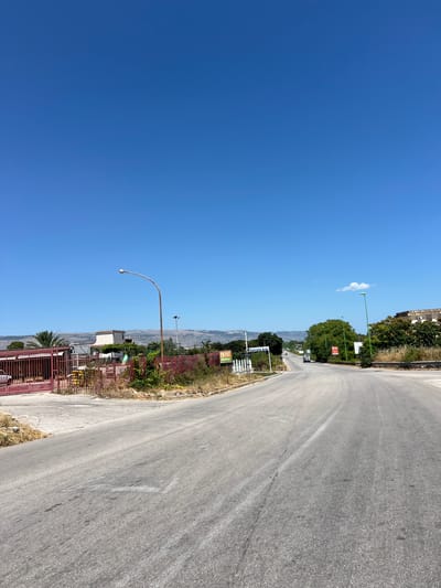 The image shows a two-lane asphalt road curving to the right, with dry vegetation and sparse trees lining its sides. A red metal gate and fence are visible on the left, with a building behind it. In the distance, a chain of low hills or mountains forms the horizon under a clear blue sky with a single white cloud. Several streetlights are positioned along the road, and various signs are placed on the right side. A white vehicle is seen in the distance on the road. The scene appears to be in an open area, possibly on the outskirts of a town or city.