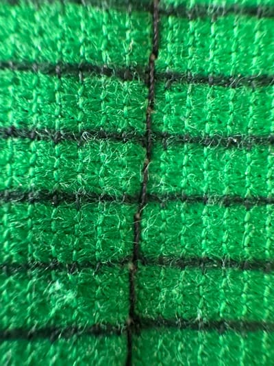 The image displays an extreme close-up of a textile, observed in Kurjan, Uzbekistan. The fabric features a prominent bright green base with a discernible grid pattern formed by dark, likely black, lines. Numerous short, fuzzy fibers constitute the green areas, giving the material a textured, possibly short-napped or looped pile appearance. A distinct, thicker vertical dark line, appearing as a seam or tightly woven structural element, runs vertically through the center of the frame. Thinner horizontal dark lines are interspersed across the green fabric, creating a checkered design. The individual threads and weave structure are clearly visible under magnification. No people, actions, or additional environmental context are present within the frame.