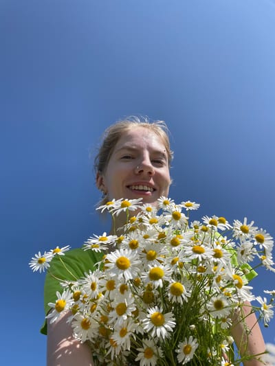 In the city of Сям-Можга, Russia, a young woman with blonde hair, wearing a bright green shirt, smiles while holding a large bouquet of white daisies with yellow centers. She has a nose ring and several earrings. The flowers are in the foreground, obscuring much of her body. The background is a clear, bright blue sky, indicating an outdoor setting on a sunny day. The image is captured from a low angle, looking upwards at the woman and the sky.