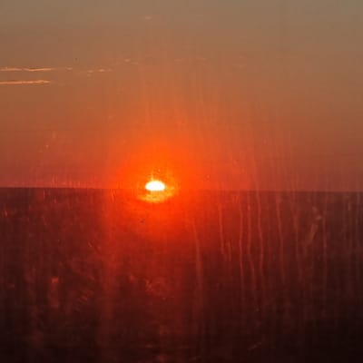 The image shows a sunset over a landscape. The sun is a bright orange sphere, partially obscured by a thin layer of clouds. The sky is a gradient of orange and yellow, fading into a darker hue at the top. The horizon line is a dark, flat band, suggesting a flat terrain such as plains or fields. The foreground is also dark, with vertical streaks that could be trees, tall grass, or a fence. There are some white specks visible in the sky and on the surface of what appears to be a window, indicating dust, dirt, or imperfections on a lens or viewing surface. The overall impression is of a natural scene viewed from indoors. The location of Бекбауыл ауылы, Kazakhstan, is not visually discernible from the image itself, as there are no identifying landmarks or features.