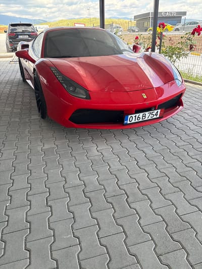 The image shows a red Ferrari with Bulgarian license plate 016 B 754 parked on a gray paved surface. In the background, there is a building with "G-AUTO" signage. Further back, there's a row of cars including a black SUV with the license plate E 4449 HK. The location is assumed to be in or near Barakovo, Bulgaria. A flower pot is visible near the building. The photo is taken at ground level.