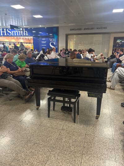 The image shows an airport scene in Luqa, Malta. A black piano is positioned in the foreground amidst a crowd of people, many of whom are seated on chairs. Behind the crowd, signage indicates the presence of a WHSmith store with advertisements for books, magazines, drinks, chocolates, sweets, and travel items. Above the store, a closed storefront displays the words "SUNGLASSES - JEWELLERY - WATCHES". The floor is tiled, and the lighting appears to be typical of an airport terminal. People are waiting around.