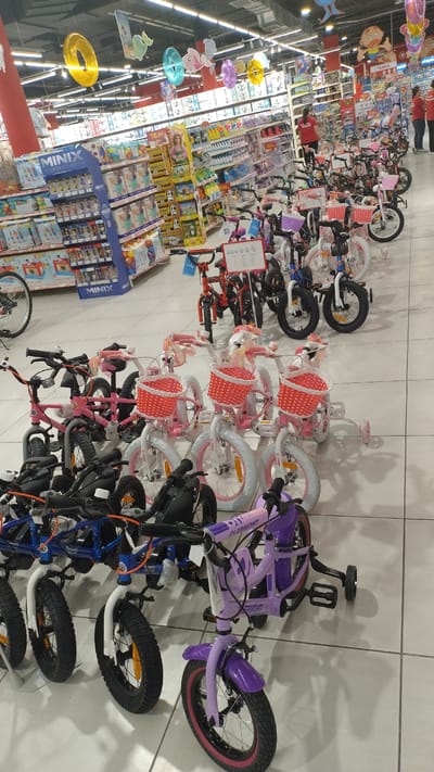 The image shows an interior view of a large retail store, possibly a department store or a toy store, located in Pylaia Municipal Unit, Greece. The foreground and midground are filled with a display of children's bicycles of various sizes and colors, predominantly pink and purple, with some white and blue models visible. Many of the smaller bikes are equipped with training wheels and front baskets, some decorated with ribbons.

In the background, shelves stocked with toys and other merchandise are visible. There are also some adult shoppers and store employees in red shirts browsing the aisles or standing near the shelves. The environment is well-lit with overhead fluorescent lighting. Decorative elements like colorful inflatable rings and cartoon character cutouts are hanging from the ceiling, suggesting a festive or child-oriented atmosphere. The floor is tiled in a light color. The overall impression is a well-organized retail space dedicated to children's products.