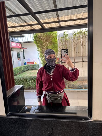 The image displays a mirror selfie of an individual located in Ban Huai Phlap, Thailand. The person, wearing a grey camouflage-patterned balaclava that obscures most of their face except for the eyes, is visible from the waist up. They are dressed in a maroon bomber jacket with white and dark blue striped cuffs and collar, over a white inner garment displaying a partial red "M". A grey fanny pack with white zippers is strapped around their waist, and a smartwatch is visible on their left wrist. The individual holds a light-colored smartphone with a rugged case in their right hand, pointed towards the mirror to capture the reflection.

The mirror itself is black-framed and appears mounted above a dark, possibly granite, counter surface, which exhibits splashes of water. Reflected in the mirror is an outdoor commercial setting. To the left of the reflection, a building features a red sign displaying the reversed text "FIVE STAR". A neatly trimmed green hedge runs across the mid-ground, beyond which slender trees with green foliage are visible. An overhead structure composed of corrugated metal roofing panels supported by dark beams is also reflected in the upper portion of the mirror. The scene is illuminated by natural daylight.