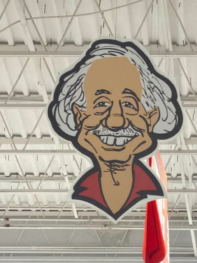 This is a photograph of a cartoon caricature of a man hanging from a ceiling. The caricature has a tan face, white hair with black outlines, a black mustache, and is wearing a red shirt with a black collar. The man's eyes are closed, and he is smiling with his mouth open, showing white teeth. The caricature is attached to the ceiling by wires, and it appears to be made of cardboard or a similar material. The background consists of a white ceiling with exposed metal beams and wires. To the right of the caricature, a large red banner or flag is also hanging from the ceiling. The scene appears to be indoors, possibly in a store or public building. The exact location in Elizabeth City, United States of America, is not specified in the image.