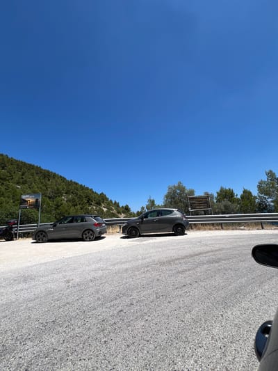 On a sunny day, two gray cars are parked on the shoulder of a road that curves along a densely wooded hillside. The road surface is asphalt with visible gravel. To the left, a dark-colored motorcycle is parked next to a sign displaying an image of rock formations and text that reads "Faraglioni." Further down the road, to the right, another sign is visible with text in Italian and English, indicating "Parco Nazionale del Gargano" and "Welcome." The hillside is covered in a thick growth of green trees, and the sky is a clear, vibrant blue. The scene appears to be in or near Mattinata, Italy, given the mention of the Gargano National Park and the Faraglioni rock formations, which are a notable feature of the Mattinata coastline.