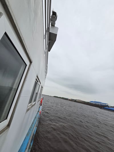 The image presents a tilted, dynamic view from the side of a boat navigating a wide body of water, likely a river, under an overcast sky. The white hull or superstructure of the boat dominates the left side of the frame, featuring several rectangular windows with dark, reflective glass. Above the windows, a blue stripe runs along the boat's exterior, from which two bright orange lifebuoys are conspicuously hung, serving as safety equipment.

On the upper deck or roof of the boat, a metallic structure is visible, including what appears to be a spotlight or navigation light and other equipment, possibly an antenna or part of a small mast. The design of the boat suggests it could be a passenger ferry or a river cruiser.

To the right, the dark, murky water stretches out, marked by ripples and small waves, indicating movement from the boat or natural current. Along the distant horizon, a strip of land is faintly visible, dotted with structures. One prominent building on the shoreline appears to have a long, blue roof or tarp covering, suggesting industrial or port facilities.

The sky is a uniform, light grey, completely overcast, implying a cloudy day without direct sunlight. The overall atmosphere is calm and subdued. The tilted perspective adds an immersive feeling, as if looking out from the moving vessel. No visible text is present in the image. Based on the provided context, the scene is likely near Nizhneleninskoe, Russia, possibly on the Amur River.