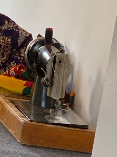 The image shows a close-up view of an antique sewing machine, predominantly black with silver accents. The machine is housed in a wooden base and is positioned on a gray, textured surface, likely a carpet. To the left of the sewing machine, a piece of fabric with a dark purple background and an intricate brown floral pattern is draped. Adjacent to this fabric is a bundle of brightly colored fabrics, including yellow, red, and green. The sewing machine itself features a needle, presser foot, and a spool pin with a red spool. The background consists of a plain white wall. The image is an indoor shot with natural lighting. No people are visible in the frame. The image does not contain any discernible elements that would allow for the determination of its location within the city of Arbagish, Uzbekistan.