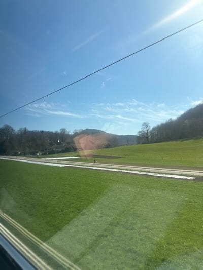 The image captures a rural landscape near Sissach, Switzerland, observed through a window displaying reflections, likely from a moving vehicle. The upper half of the frame is dominated by a clear, bright blue sky containing scattered, faint cirrus clouds. Intense sun glare emanates from the upper right, and a single, thin, dark utility wire stretches horizontally across the sky.

The lower half of the image features vibrant green grassy fields. A paved road runs horizontally across the midground. Parallel to the road, on the side closer to the viewer, is a continuous strip of white material, consistent with an agricultural covering or remaining snow. A lone individual, dressed in dark attire, is visible riding a bicycle from left to right on this road.

In the background, a cluster of leafless deciduous trees and several structures, including buildings with visible roofs, are present on the left. A large, tree-covered hill, primarily composed of bare deciduous trees, rises steeply from the right side of the frame towards the center. The environment suggests late winter or early spring conditions, characterized by green fields and dormant trees under clear daylight.
