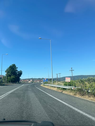 This is a photograph taken from the perspective of someone in a vehicle, looking forward along a paved road in Νέα Μεσολακκιά, Greece. The road has double white lines marking the lanes and appears to be a highway or a major thoroughfare. On the right side of the road, there is a metallic guardrail with dry grass and vegetation growing alongside it. Beyond the guardrail, there are some trees and bushes, and further in the background, there are rolling hills with scattered houses and red-tiled roofs. A small sign with "50" in a blue circle, indicating a speed limit, is visible on the right side of the road, along with a blue directional sign. On the left side of the road, there are more trees, including a large, dense one, and streetlights. Overhead, the sky is a clear, bright blue with a few wispy clouds. There are also several utility poles with wires visible. A red and white sign with Greek text is also present on the right side of the road. The overall scene depicts a sunny day on a road leading towards a town or village.