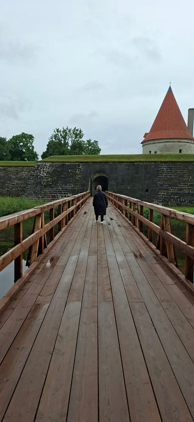 The image depicts a person walking across a wooden bridge towards a historical stone fortress in Kuressaare, Estonia. The fortress, likely Kuressaare Castle, is visible in the background with its distinctive red conical tower. The bridge is constructed with wooden planks and railings, and it spans over a body of water, possibly a moat. The fortress wall, made of large dark stones, features an arched entrance. Lush green grass and trees surround the fortress. The sky is overcast, suggesting a cloudy or rainy day. The person, seen from behind, is wearing a dark, quilted coat and dark trousers. The overall scene conveys a sense of historical exploration and a slightly somber atmosphere due to the weather.