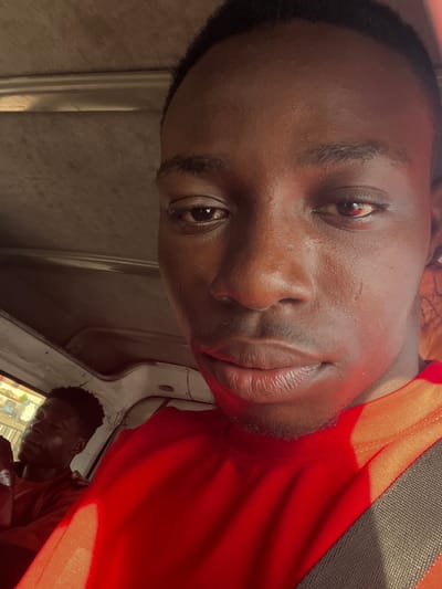 The image is a close-up selfie of a Black male, taken inside what appears to be the cabin of a vehicle. The man is looking directly at the camera, with his face filling most of the frame. He has dark skin, short black hair, and is wearing a bright red shirt. The lighting suggests it is daytime. In the background, slightly out of focus, another Black male is visible, leaning back and appearing to be asleep or resting. This individual is also wearing a red shirt. The interior of the vehicle is visible, with a beige-colored ceiling and interior paneling. A hint of the outside environment, possibly urban, can be seen through a window on the left. The image is taken in Okwe, Nigeria.