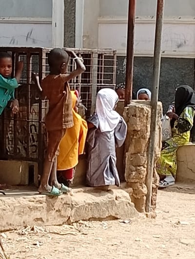 This outdoor scene, set in Fanisau, Nigeria, captures a moment of daily life featuring several children and adults under bright daylight. In the foreground, a young boy with dark skin, wearing a brown shirt and light green sandals, stands with his back to the viewer on a rough, raised stone step. His arms are raised, seemingly adjusting something on his head or hair, which is adorned with a white bracelet. To his left, another young boy in a green shirt peeks out from behind a metal crate, looking directly at the camera.
Further to the right, a child in a grey, flowing robe and white head covering stands with their back to the camera, partially obscured by a sturdy stone pillar. Between the two central children, a glimpse of a child in vibrant yellow or orange clothing is visible. Behind the stone pillar, two sets of eyes can be seen peeking out, suggesting other individuals are observing the scene. On the far right, an adult woman with a black head covering and a colorful patterned dress (green, yellow, and black) is seated on a low stone ledge, wearing white sandals.
The background features light-colored building walls with window openings covered by metal grates. The ground beneath the stone steps appears dusty or unpaved. The bright, direct sunlight indicates a sunny, dry day. There is no visible text in the image.