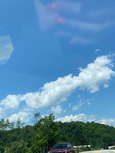 The image shows a bright, sunny day with a blue sky and scattered white clouds. The scene appears to be a rural or suburban area, possibly near a highway, given the presence of a road and vehicles. The landscape is characterized by lush green trees and rolling hills. In the foreground, a red SUV is visible on the right side of the road. Further down the road, another black vehicle can be seen. The overall impression is one of a scenic drive on a clear day. The prompt mentions the city of Ripon, Canada, which is not visually identifiable in the image itself.
