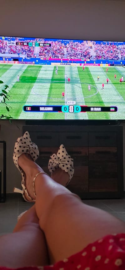 A television screen displays a soccer match between Belgium and Iran, with the score at 0-0 in the 67th minute. The game is taking place in Los Angeles. In the foreground, a person's legs are visible, wearing white polka-dot heels. The person is seated, with their legs crossed and resting on a piece of furniture. The room appears to be a living area, with a plant visible in the background. The overall scene suggests a casual setting, possibly a home, where the person is enjoying watching the soccer game. The location is not Lieli, Switzerland.
