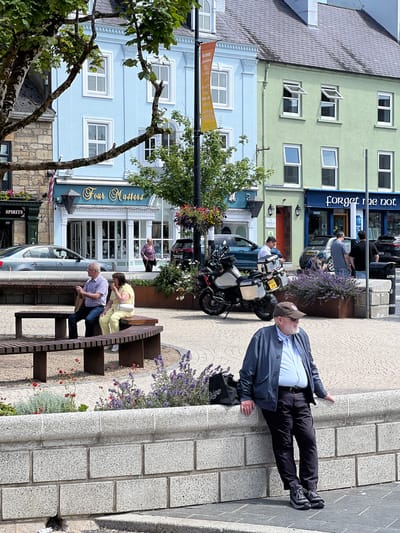 The image depicts a street scene in Donegal Town, Ireland, likely during daytime. In the foreground, a man wearing a blue jacket, striped shirt, dark trousers, and dark sneakers leans against a low stone wall. To his left, a black bag is placed on the wall near a cluster of purple flowers. In the midground, a couple sits on a wooden bench. The man wears a checkered shirt and dark trousers, while the woman wears a yellow outfit and a white handbag. Behind them, a street with several buildings is visible. The buildings are painted in pastel colors: a pale blue building on the left with a sign that reads "Four Masters," and a light green building to its right with a sign reading "forget me not." A motorcycle is parked on the street. In the background, more buildings and trees can be seen, along with parked cars. The overall scene suggests a relaxed, urban atmosphere.