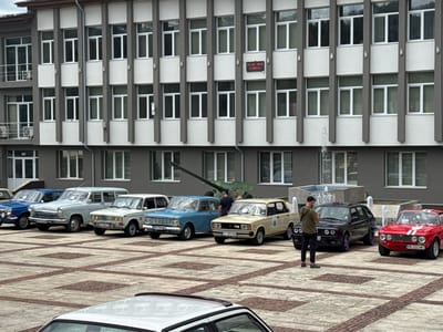 In Hadzhidimovo, Bulgaria, on June 13, 2026, a collection of vintage cars is displayed in an outdoor plaza in front of a modern, multi-story building with numerous windows. The building's facade is gray and white, with dark vertical columns. A digital clock on the building reads "13. 06. 2026." A fountain is visible in the background. Several classic cars, including a light blue sedan, a beige sedan, a light gray station wagon, and a red coupe, are parked in a grid pattern on a patterned paved surface. Two men are observing the cars, one in a green jacket and black pants, and another in a dark shirt. A military cannon is also present in the background, behind the cars.