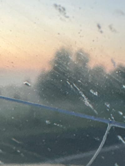 The image depicts a blurry, abstract scene with a gradient of colors suggesting a sunrise or sunset. The foreground appears to be a dirty or smudged surface, possibly a window, with streaks and water droplets. In the mid-ground, indistinct shapes of trees are visible against a soft, warm-toned sky. A thin, dark horizontal line runs across the mid-section of the image. The overall impression is one of an outdoor view through a soiled barrier, potentially captured at dawn or dusk. No specific city features or recognizable landmarks from Krapivinskiy, Russia are discernible due to the image's lack of clarity.