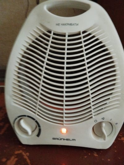 The image shows a white electric fan heater, likely in a home setting. The fan heater is the primary object, with its distinctive ribbed front grille and two rotary knobs on the lower front panel. One knob appears to be for temperature control, with markings indicating settings like "0", "1", and "2". The other knob's function is less clear from the image, but it also has a dial with markings. A small orange indicator light is illuminated on the front, suggesting the heater is operational.
Above the grille, there is some text printed on the casing. It is in Cyrillic script and appears to say "НЕ НАКРИВАТИ", which translates to "DO NOT COVER" in English, a common safety instruction for heaters. Below the indicator light, the brand name "GRÜNHELM" is visible.
The background is out of focus but suggests an indoor environment. The texture of the background hints at a wall or furniture, possibly made of wood or fabric, with a warm, neutral color palette. The lighting in the image is somewhat dim, with the orange indicator light being the brightest point. There are no people visible in the image. The overall impression is of a functional appliance ready for use. The location context is Sheptytskyi, Ukraine, which suggests this is likely a domestic setting during colder weather.