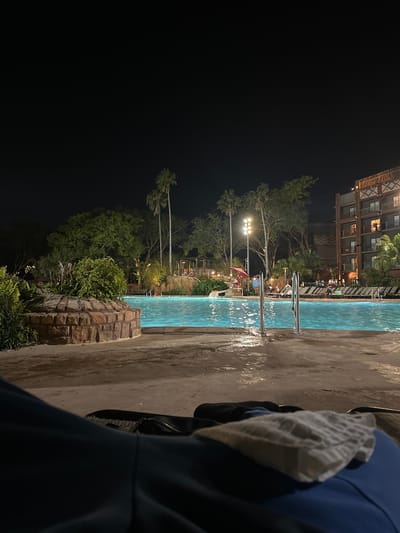 The image shows a night scene at a pool area, likely at a resort. The location is identifiable as Disney's Animal Kingdom Lodge in Bay Lake, Florida, United States, based on the distinctive architectural style of the buildings in the background. The pool is illuminated and shows reflections on the water surface. Pool ladders are visible. Trees and vegetation surround the pool area. Floodlights provide additional illumination. A structure, possibly a lifeguard stand, is present near the pool. In the foreground, a person is partially visible, wearing a blue jacket with a grey hood.